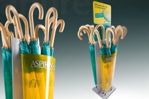 Practical umbrella stand as an action display/theme display for aspirin. The basic basis is a stable metal frame. Colored eye-catchers for customers are yellow-backprinted and branded PET-G fronts and the exchangeable cardboard crowner.